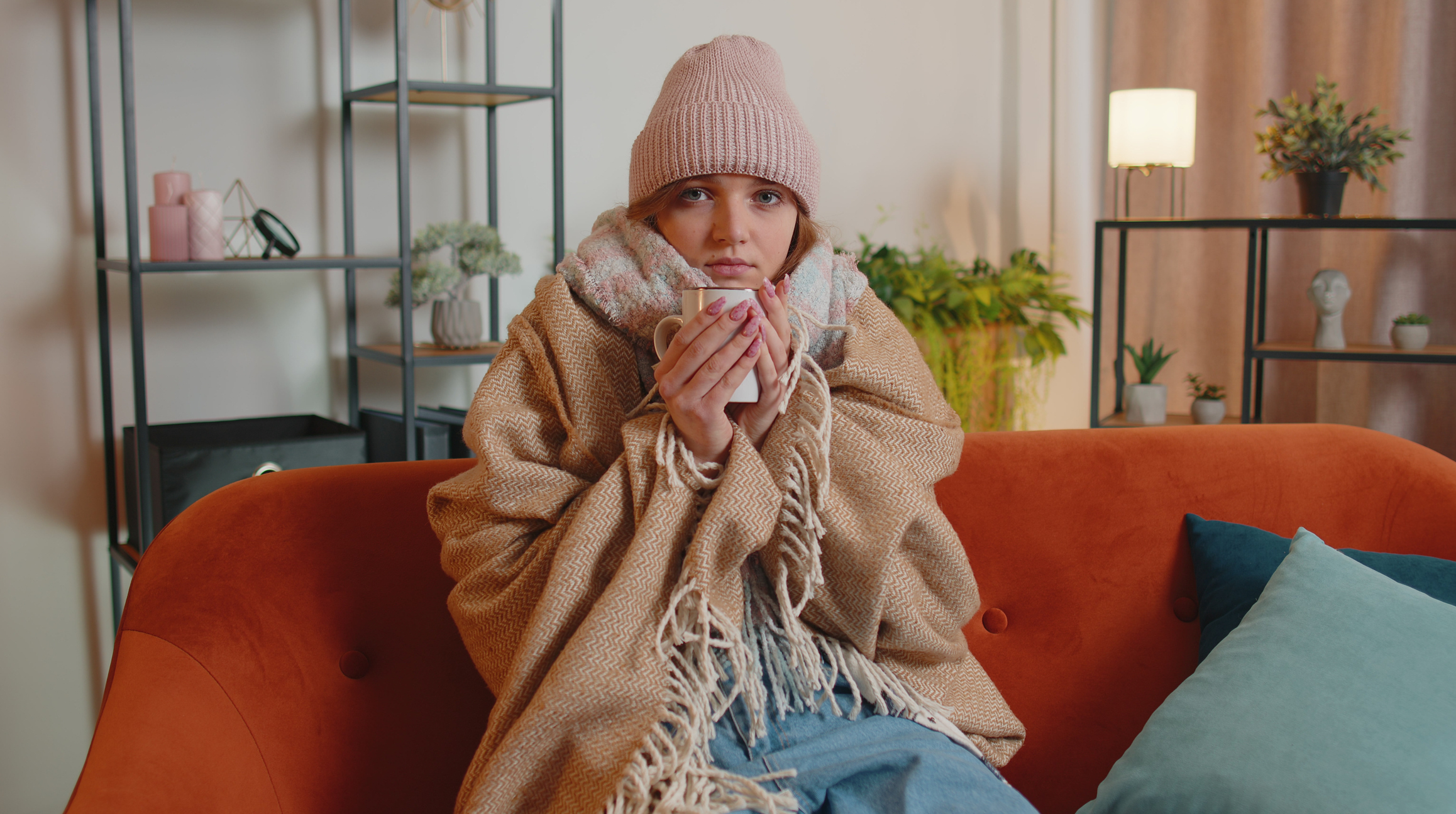 Young woman in hat shivering on sofa drinking hot tea